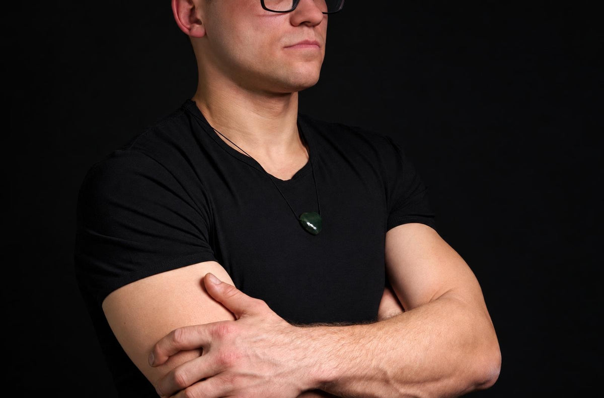 A man in a black t-shirt wears a dark green, heart-shaped pendant on a thin cord against a dark background.