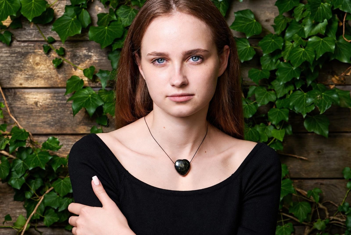 A woman wears a black top and a necklace with a dark, heart-shaped pendant against a wooden wall with green ivy.