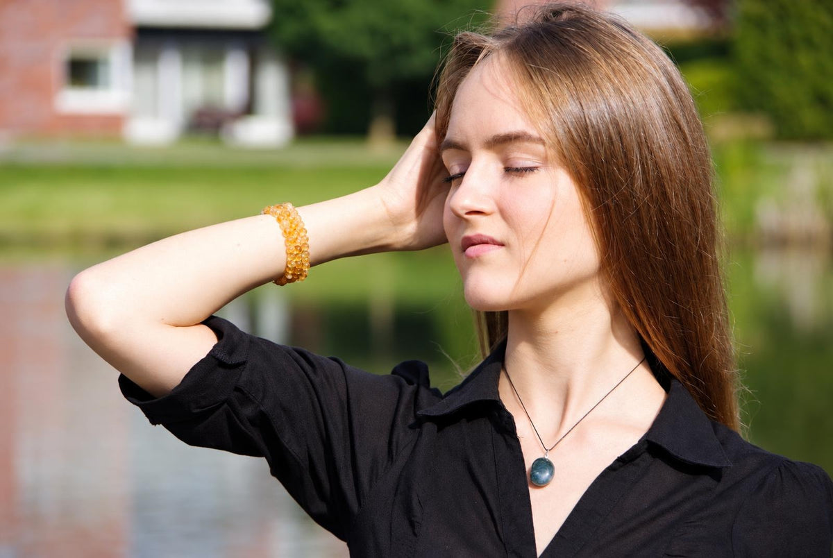 A woman wears a blue green moss agate pebble pendant on a black cord, standing outdoors with one hand behind her head.
