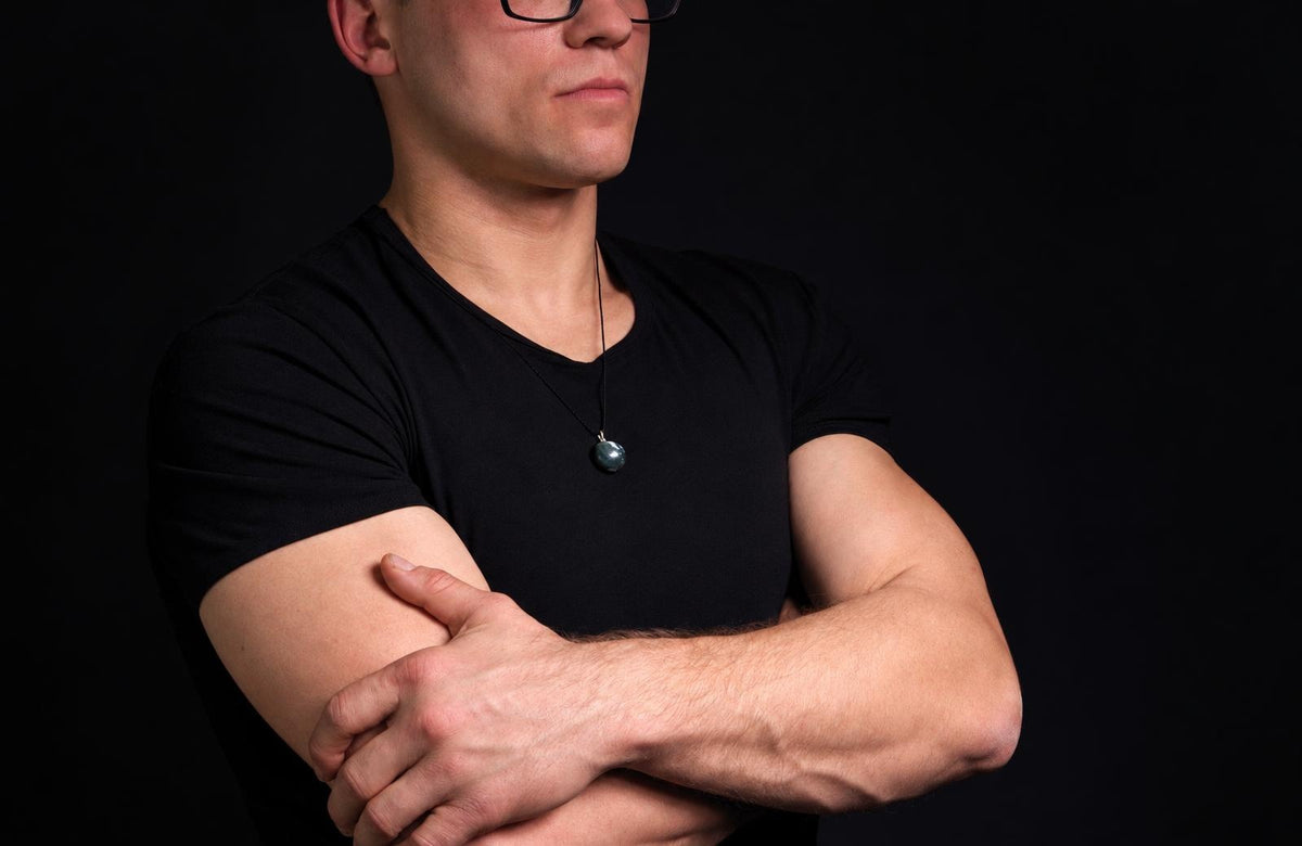 A man in a black t-shirt wears a dark green, round pendant on a thin cord against a dark background.
