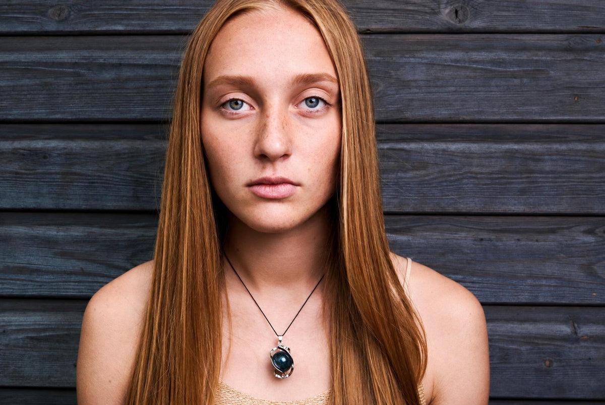 A woman with long reddish-brown hair wears a pendant with a dark blue stone in a silver-toned setting against a dark wooden background.