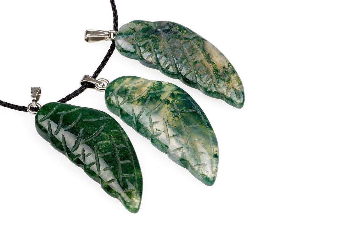 Three carved moss agate wing pendants show green branching patterns and pale translucent areas, arranged with black cords on a white background.