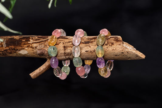 Three faceted nugget bracelets with pink, yellow, green and purple stones rest on a rough wooden branch against a dark background.