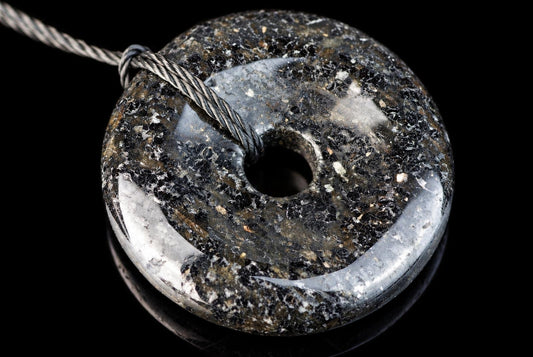 A polished, donut-shaped pendant with a dark, speckled surface and silver-toned highlights, threaded on a twisted cord against a black background.