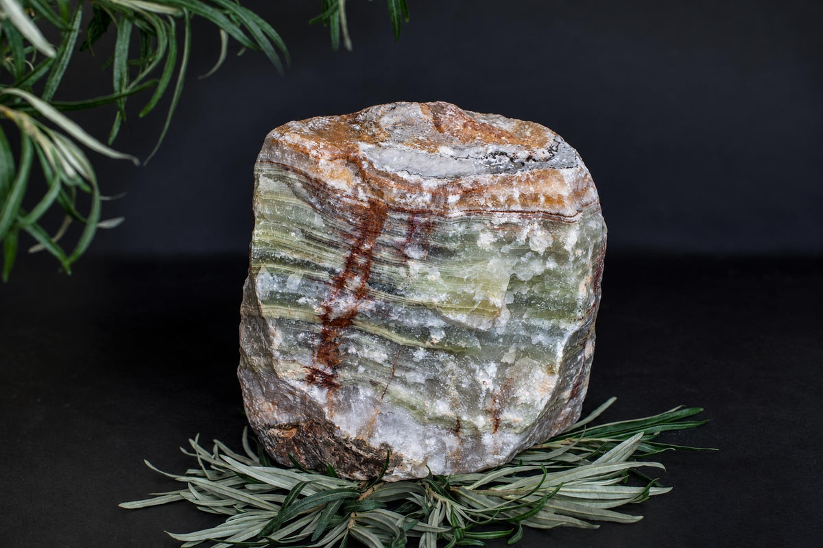 A large, rough stone with green and white horizontal layers and brown veins sits on a dark surface among rosemary sprigs.