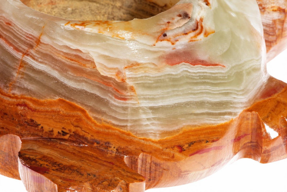 A polished onyx bowl with layered white, orange and brown bands sits on a wooden base against a pale surface.