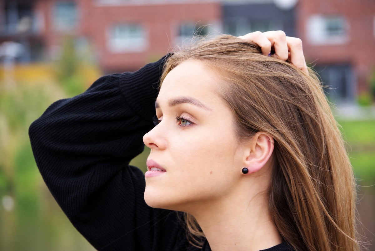 A woman with long brown hair wears a small black stud earring in her earlobe, seen from the side against a blurred outdoor background.