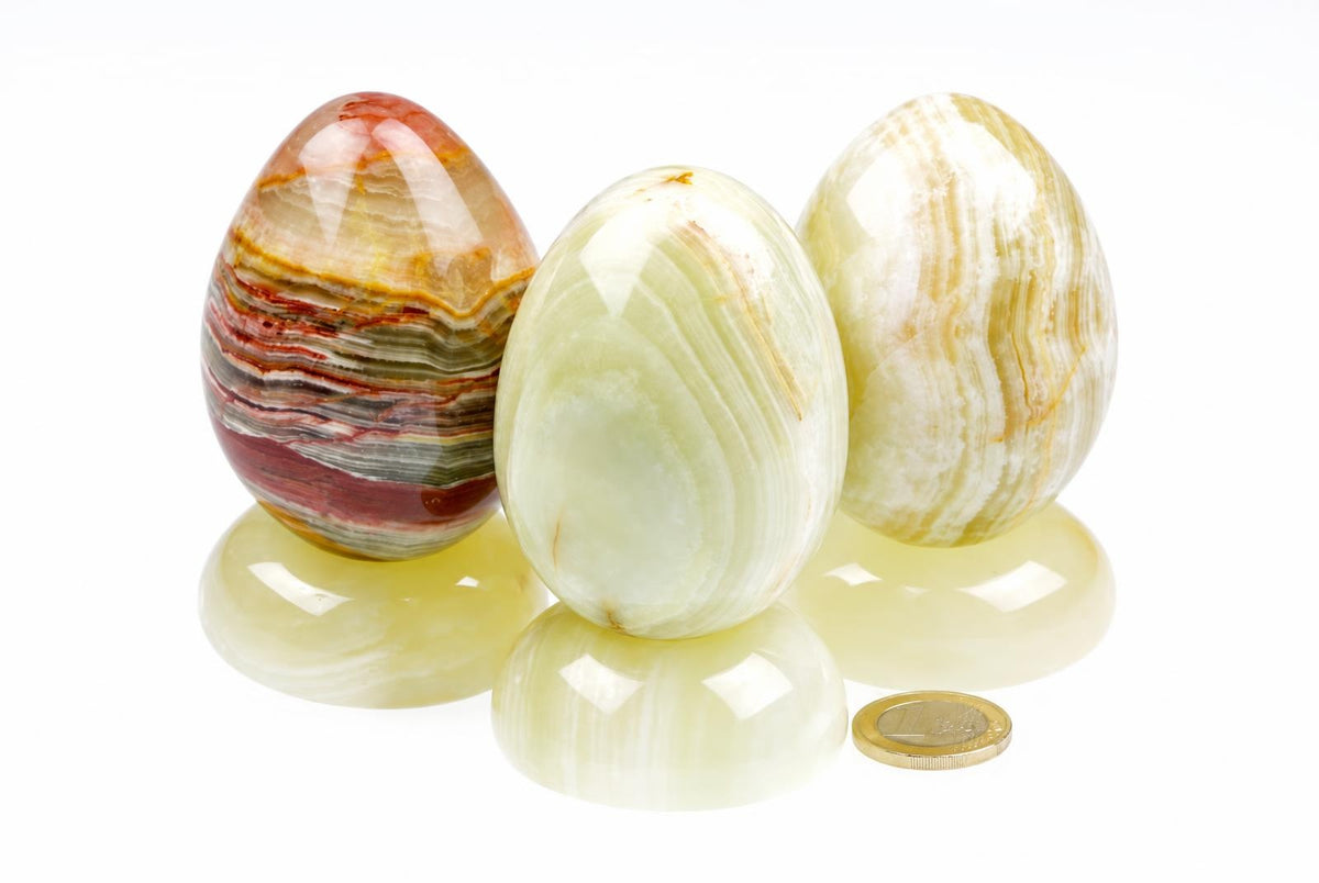 Polished onyx eggs show cream, pale green and brown bands, displayed on matching rounded stands beside a coin on a white surface.