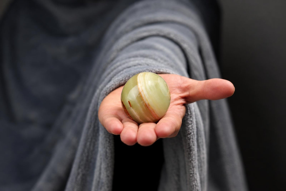 A hand holds a polished, round stone with pale green and brown stripes against a dark background.