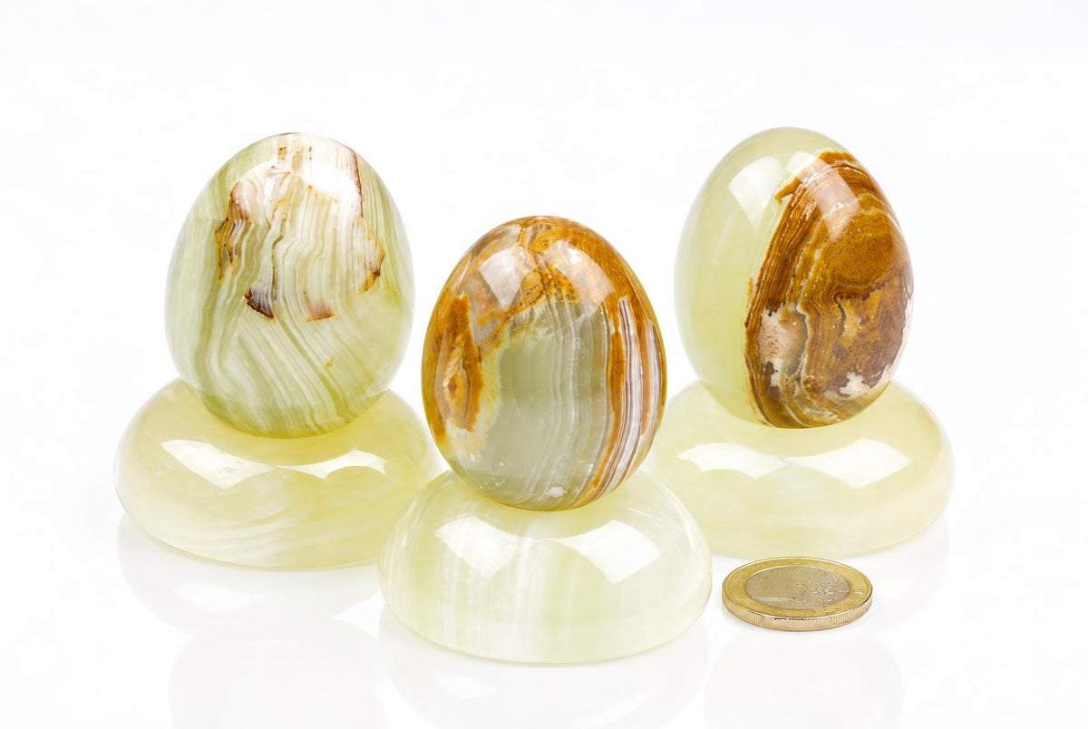Polished onyx eggs with pale green, cream and golden brown bands sit on matching rounded stands beside a coin against white.