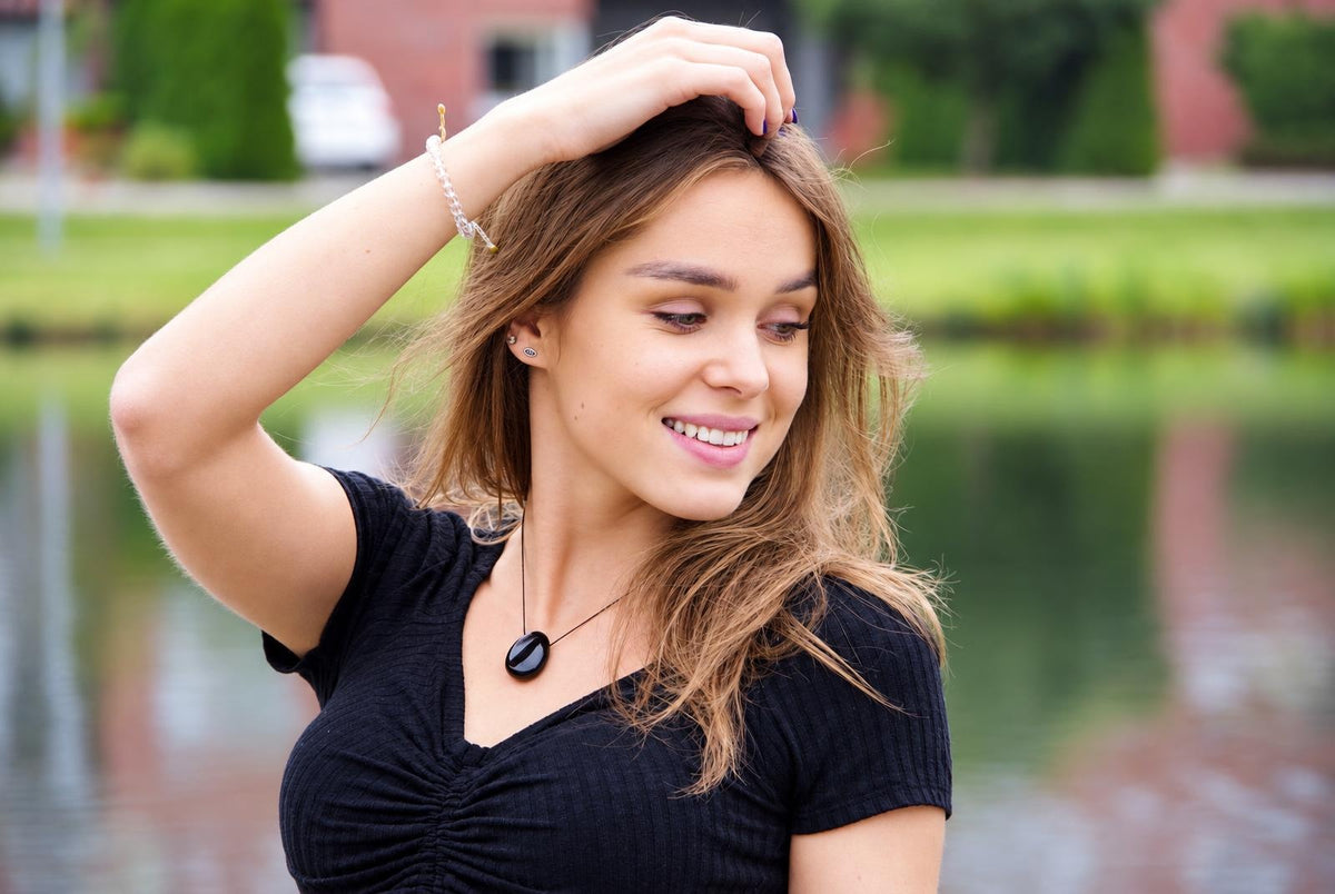 A woman wears a black top and a necklace with a dark oval pendant on a thin cord, standing outdoors near water.