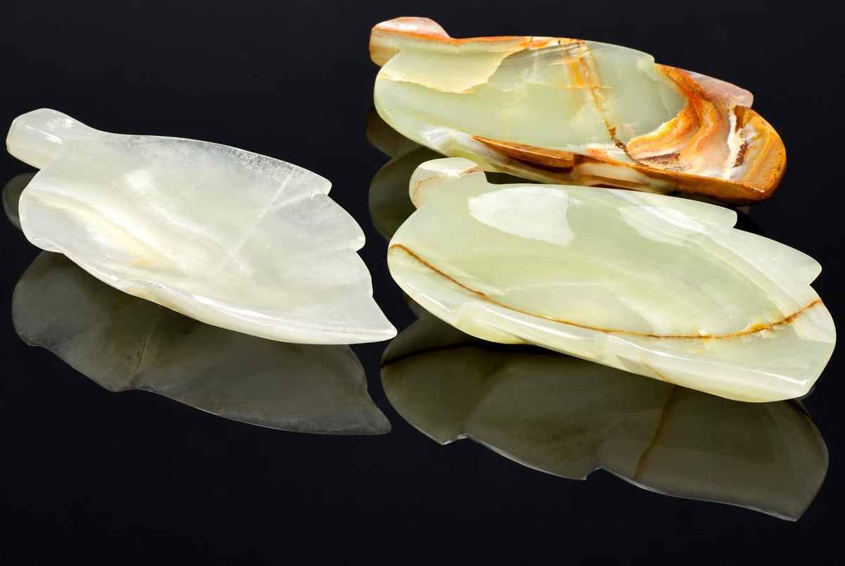 Three polished leaf-shaped plates in pale green, white and brown-toned stone on a dark reflective surface.
