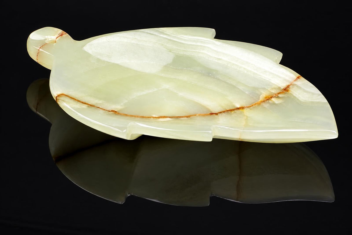 A polished, pale green onyx plate carved into a leaf shape with brown veining, resting on a dark reflective surface.