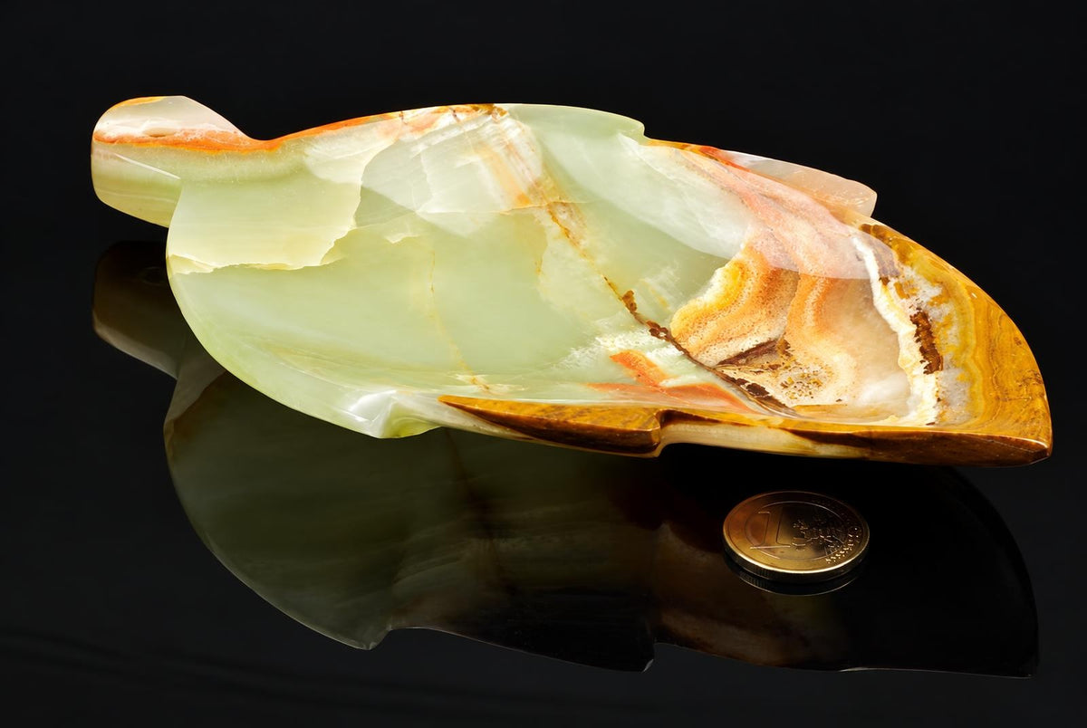 A polished onyx plate shaped like a leaf, featuring pale green and orange-brown bands, resting on a dark reflective surface with a coin nearby.