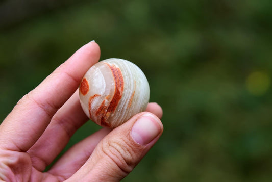 A hand holds a polished, round stone sphere with white and reddish-brown swirling bands against a blurred green background.
