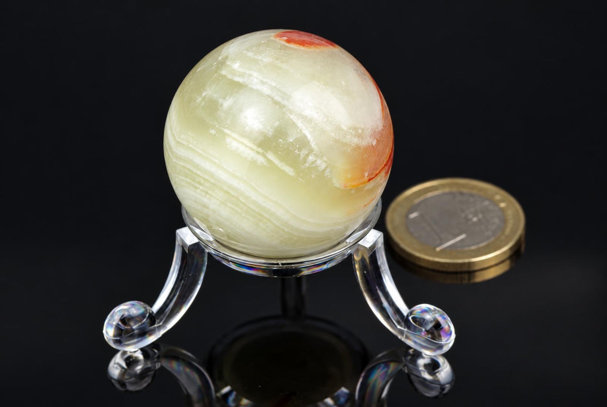 A polished, pale yellow sphere with reddish-brown bands sits on a clear acrylic stand against a dark surface.