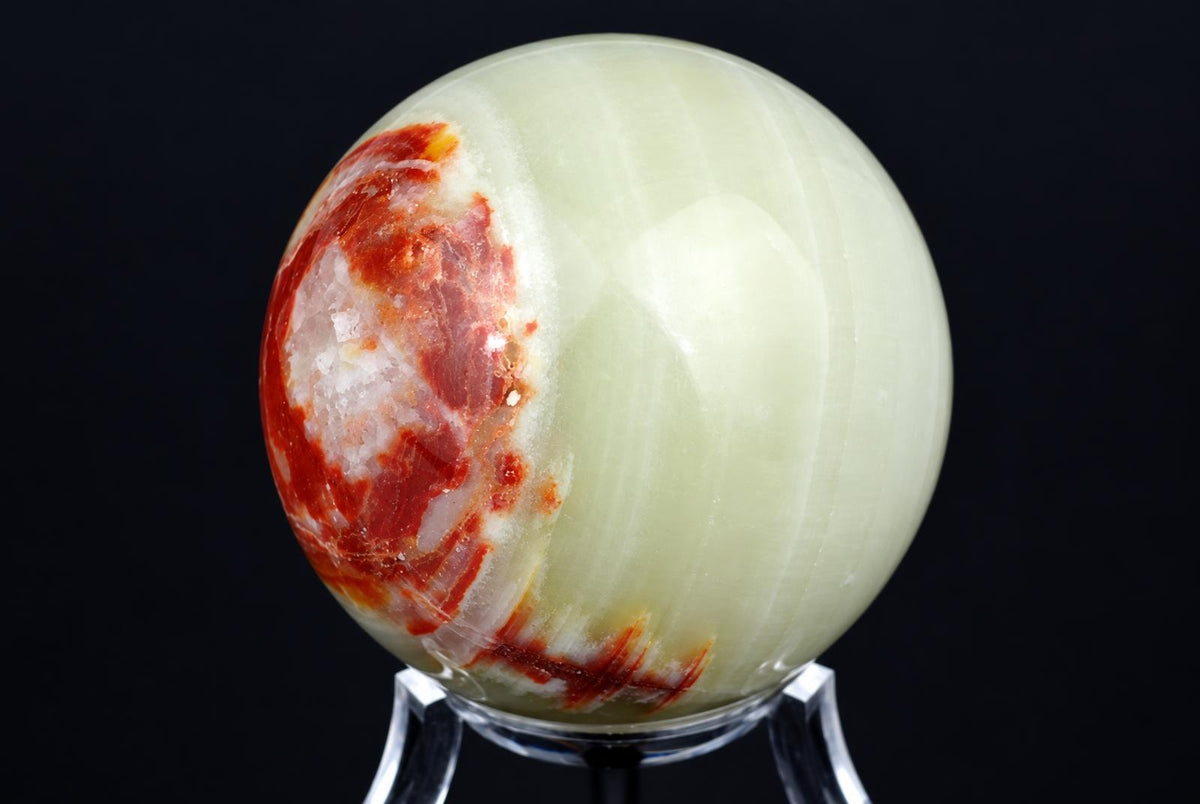 A polished, pale green sphere with red and white patches sits on a clear stand against a dark background.