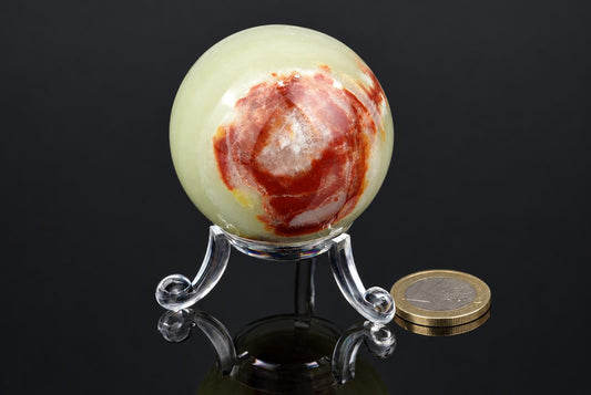A polished, pale green sphere with red and white swirls sits on a clear stand beside a coin against a dark surface.