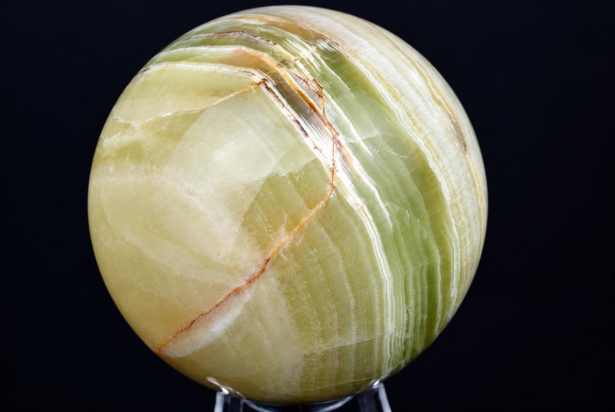 A polished, round sphere of green and white banded stone with thin brown lines on a dark surface.