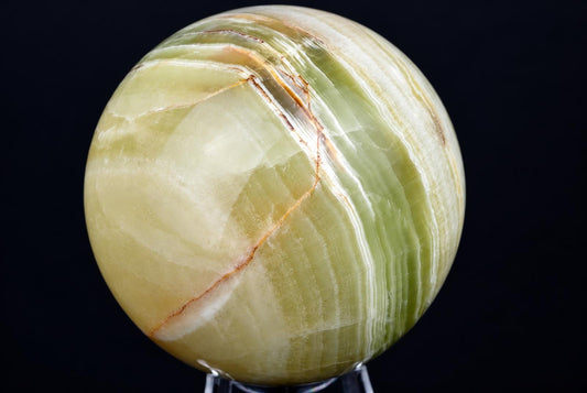 A polished, round sphere of green and white banded stone with thin brown lines on a dark surface.