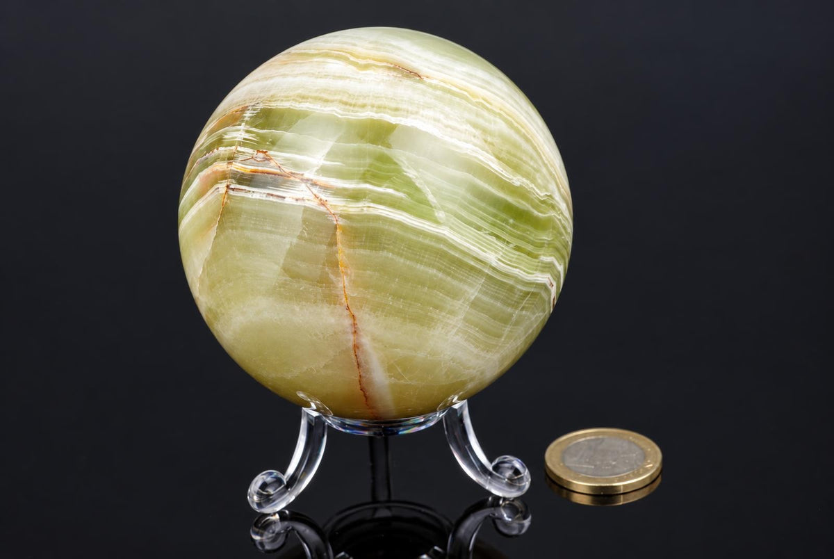 A polished green and white striped onyx sphere sits on a clear stand against a dark surface, with a coin nearby for scale.