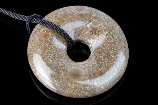 A polished, donut-shaped pendant with a central hole and fossil-like patterns in beige, brown, and white tones on a dark surface.