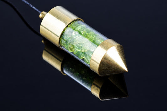 A pendulum with pale green chips inside a clear tube and gold-coloured end pieces rests on a dark reflective surface.