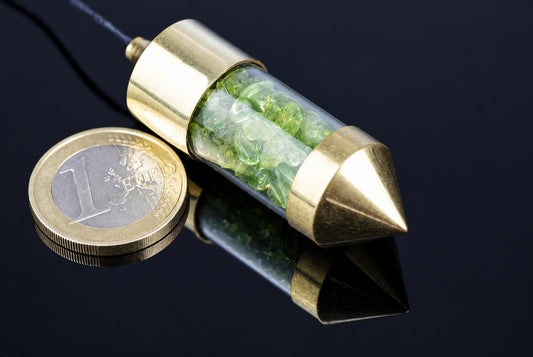 A brass pendulum with a clear tube containing green and pale stones, resting on a dark reflective surface next to a one euro coin.