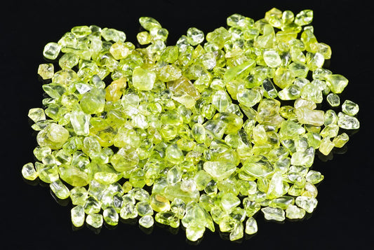 A pile of small, rough-cut peridot gemstones in lime green and yellow-green tones on a dark surface.