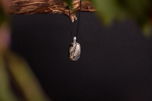 A polished, rectangular pendant with black and beige swirls hangs from a black cord on a dark surface.
