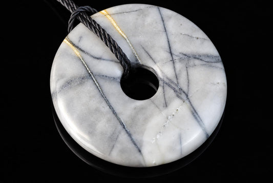 A round, white and grey veined pendant with a central hole, threaded on a dark cord against a black background.