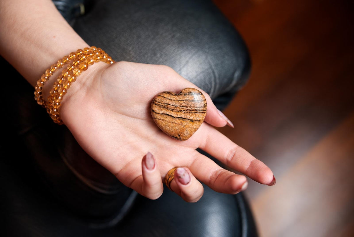 A hand holds a polished, heart-shaped stone with brown and black stripes on a dark surface.