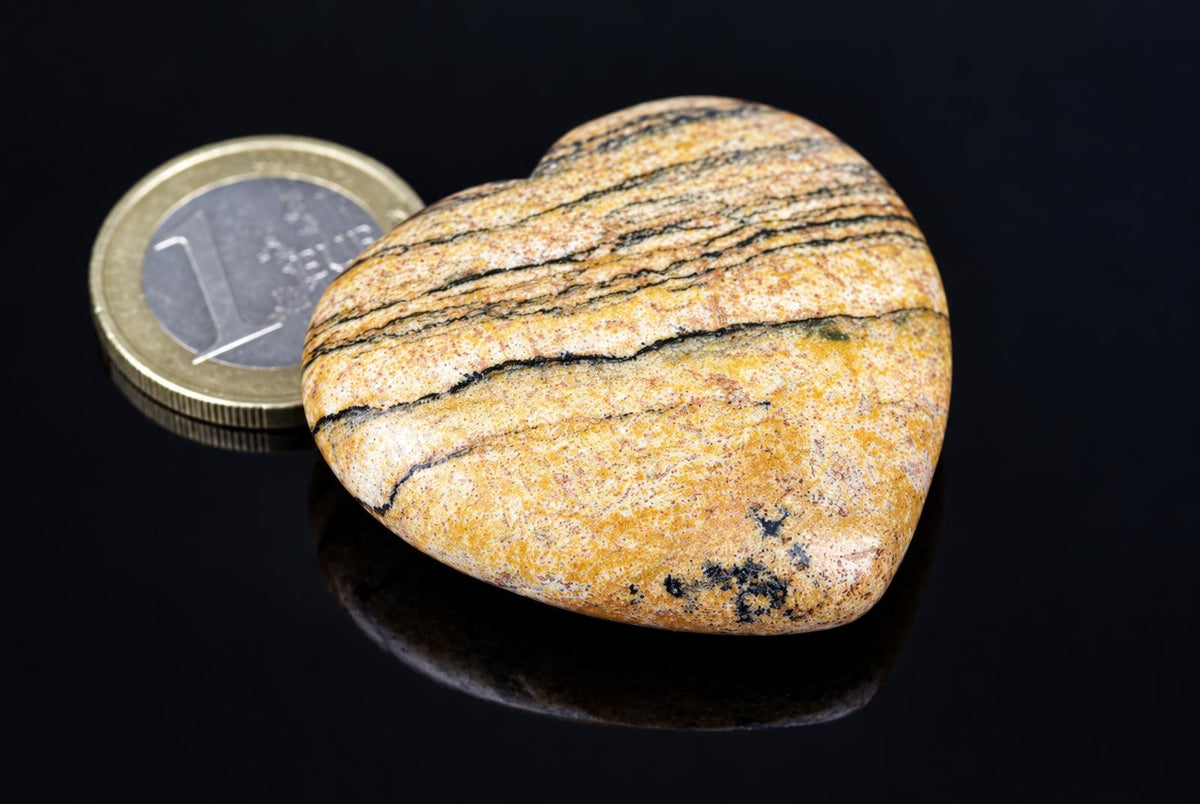 A polished, heart-shaped stone with yellow and black stripes rests on a dark surface next to a coin.