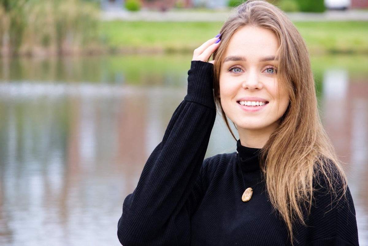 A smiling woman with long brown hair wears a black turtleneck and a light-coloured oval pendant on a dark surface.