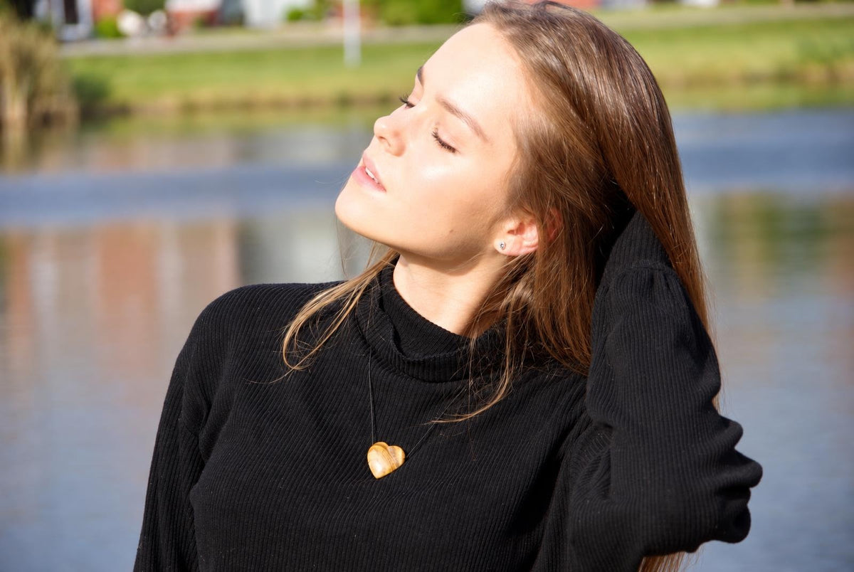 A woman wears a black ribbed sweater with a light brown heart-shaped pendant necklace against a blurred outdoor water background.