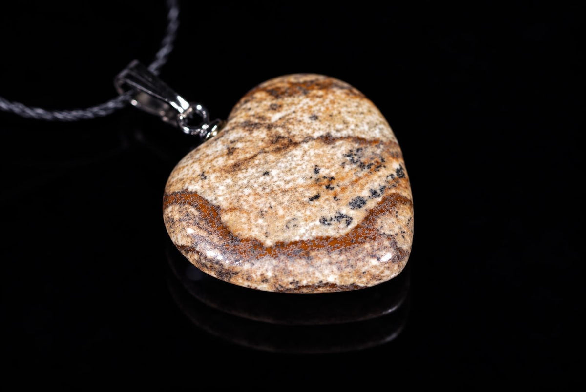 A heart-shaped jasper pendant with brown, black and cream patterns hangs from a silver-toned bail on a dark surface.