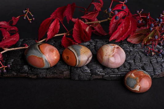 Four polished, oval-shaped stones with swirling bands of red, grey, and cream rest on a dark wooden surface beneath red leaves.