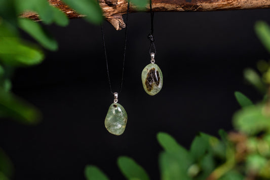 Two green prehnite pendants with dark inclusions hang from black cords on a wooden branch against a dark background.