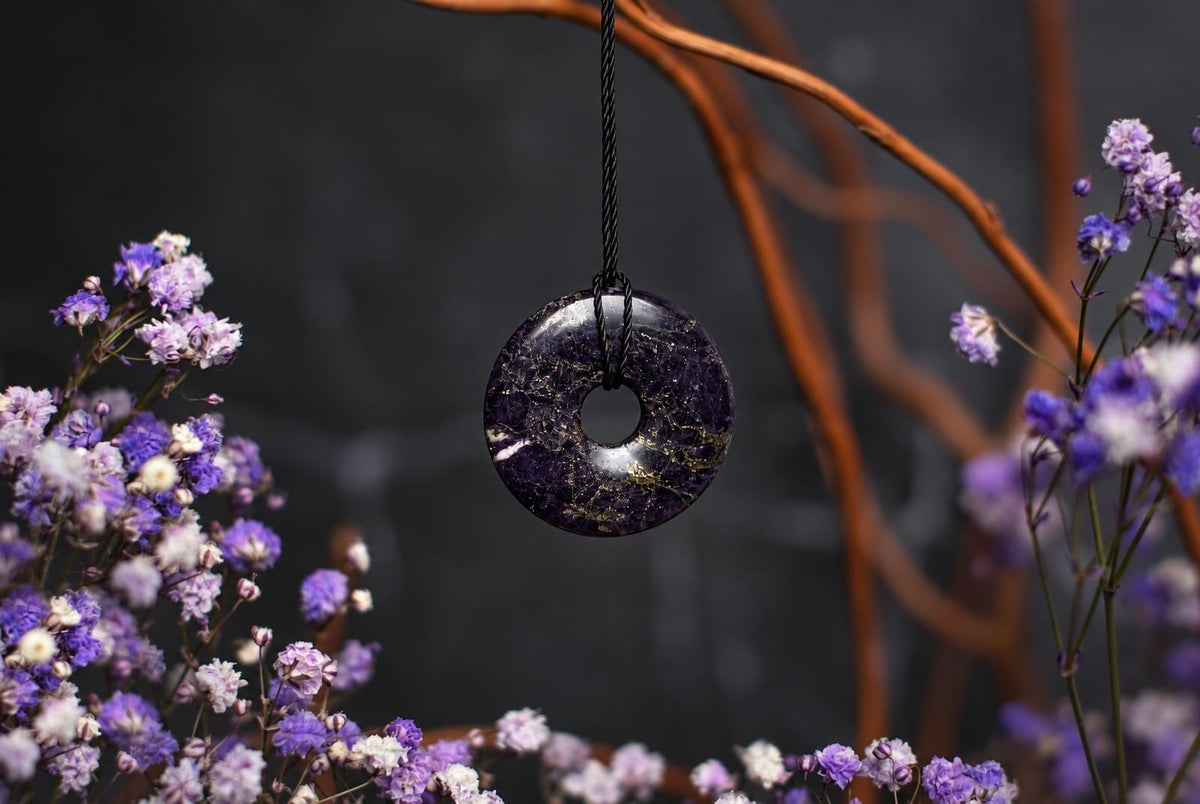 A dark purple circular stone pendant with a central hole hangs from a black cord among small purple flowers and dried stems.