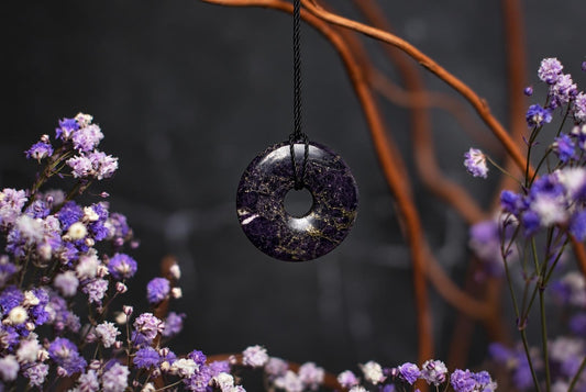 A dark purple circular stone pendant with a central hole hangs from a black cord among small purple flowers and dried stems.