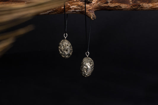 Two oval pendants with a rough, metallic gold-coloured texture hang from black cords on a dark wooden branch against a black background.