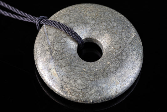 A grey, donut-shaped pendant with a cracked texture and central hole, threaded on a dark cord against a black surface.