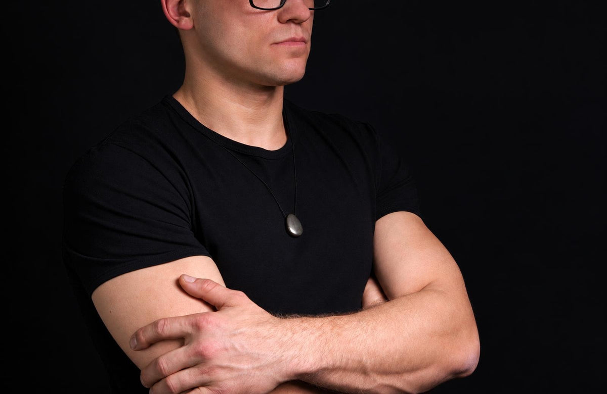 A man in a black t-shirt wears a silver-toned oval pendant on a thin cord against a dark background.