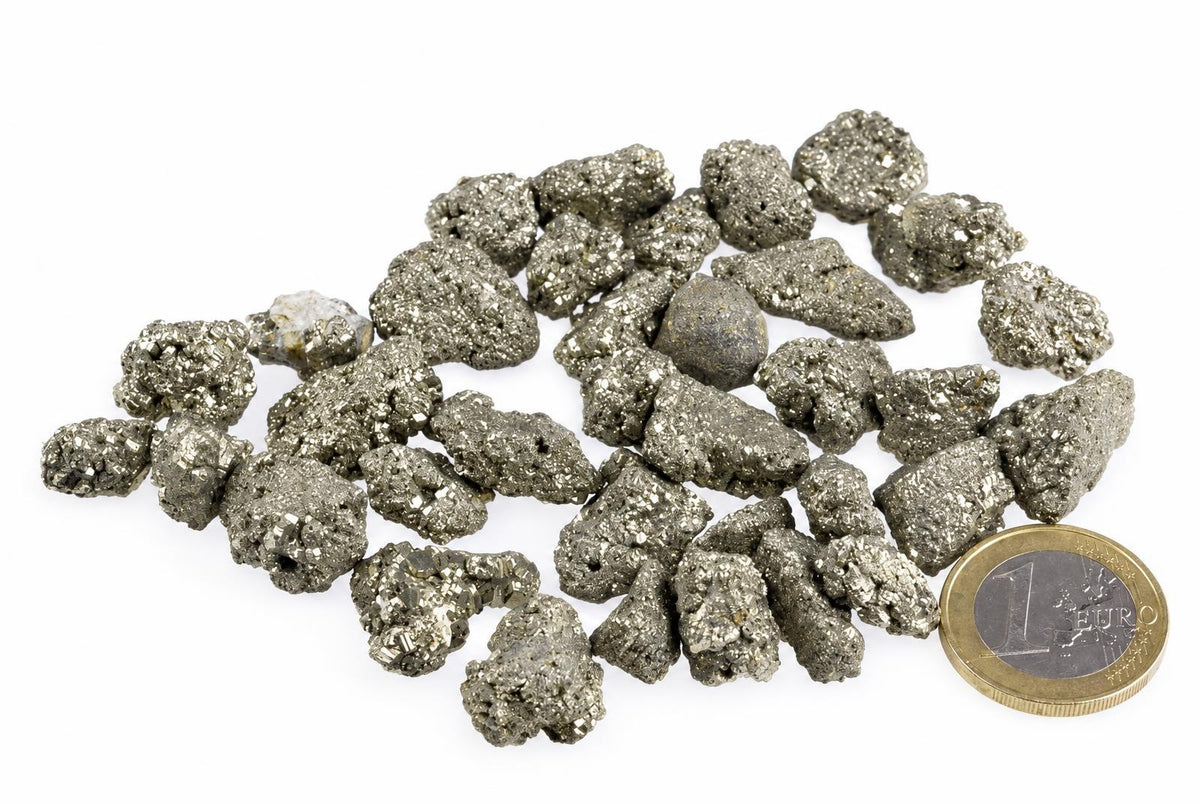 A pile of small, metallic grey and gold-coloured pyrite crystals on a white surface with a one euro coin for scale.