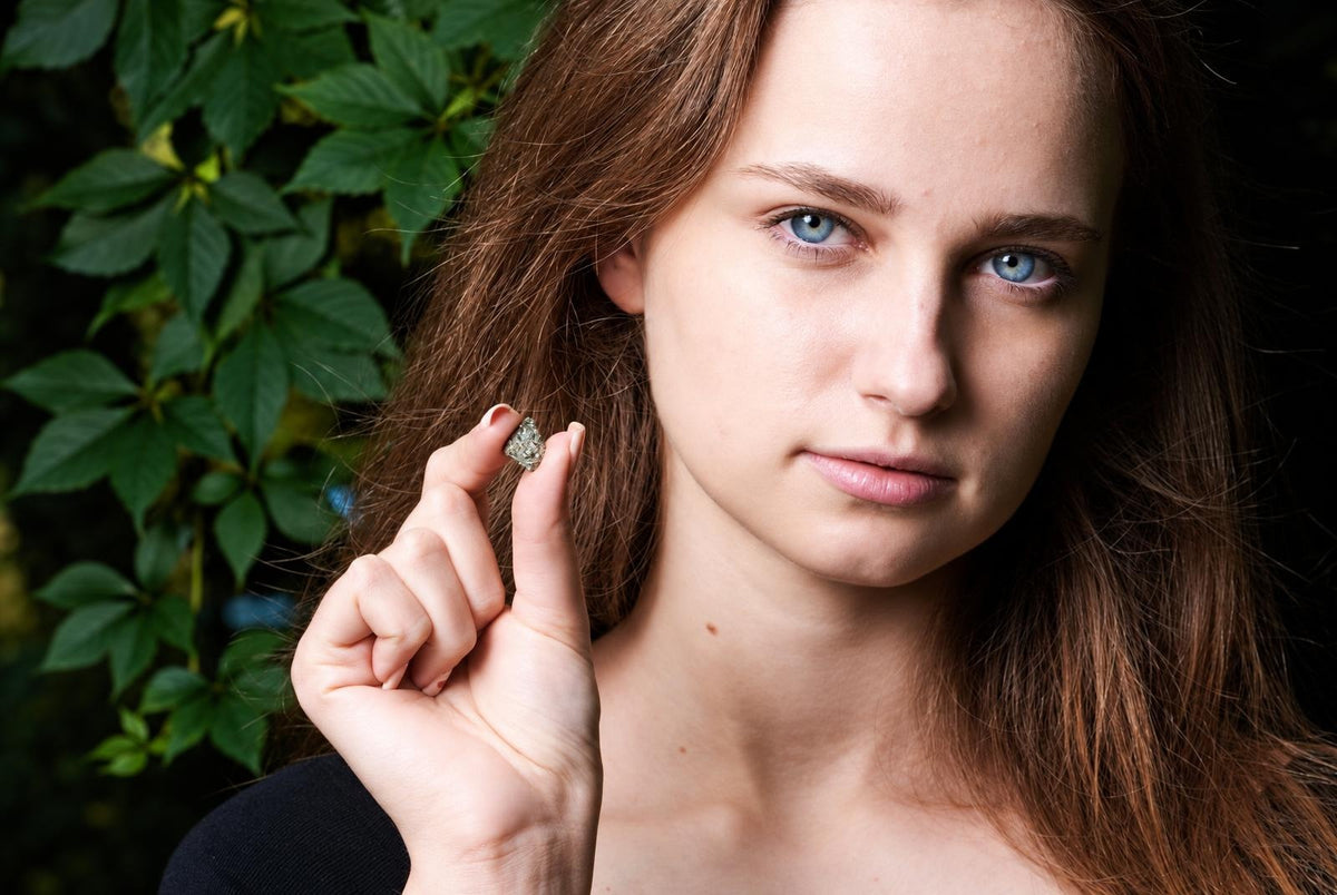 A woman holds a small, rough, metallic grey-green crystal between her thumb and forefinger against a dark surface with green leaves.