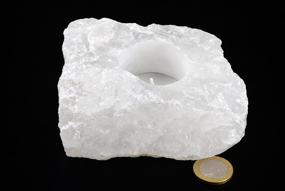 A rough white stone candle holder with a round central recess and a tealight sits beside a coin against a black background.