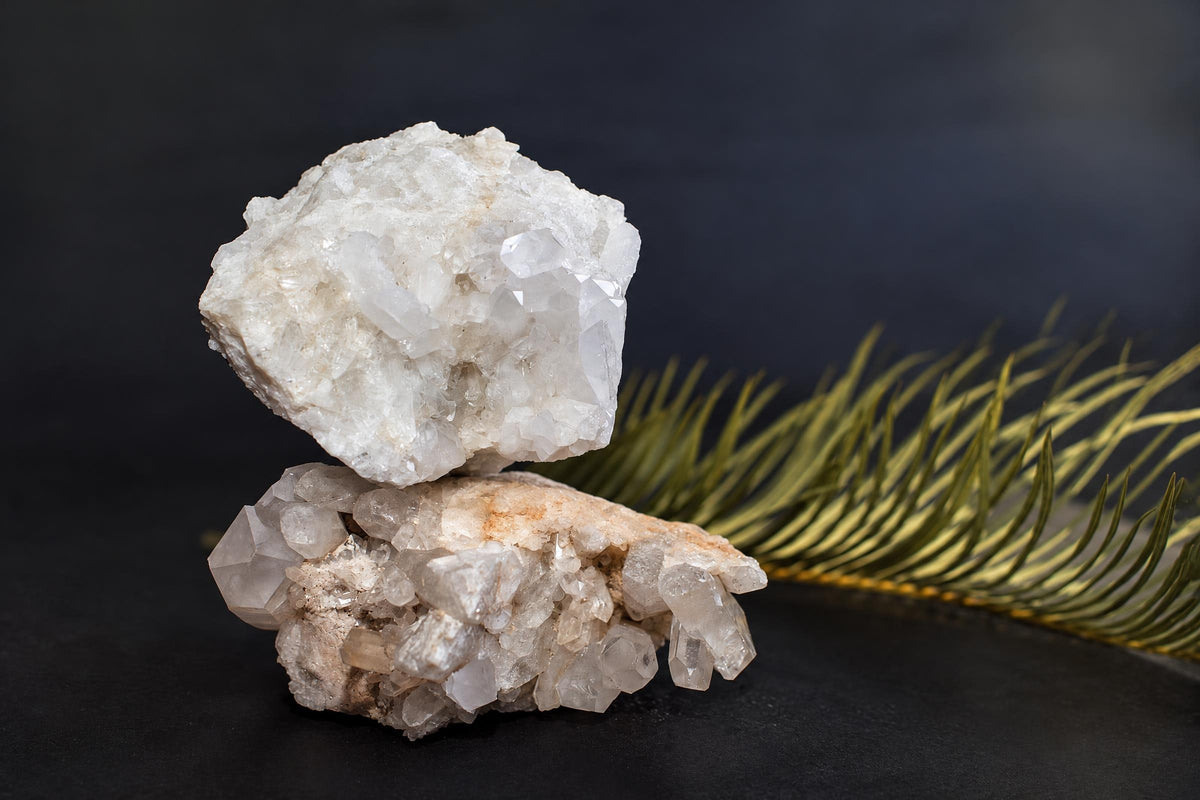Two stacked quartz crystal clusters with clear, white crystals on a dark surface next to a green palm frond.