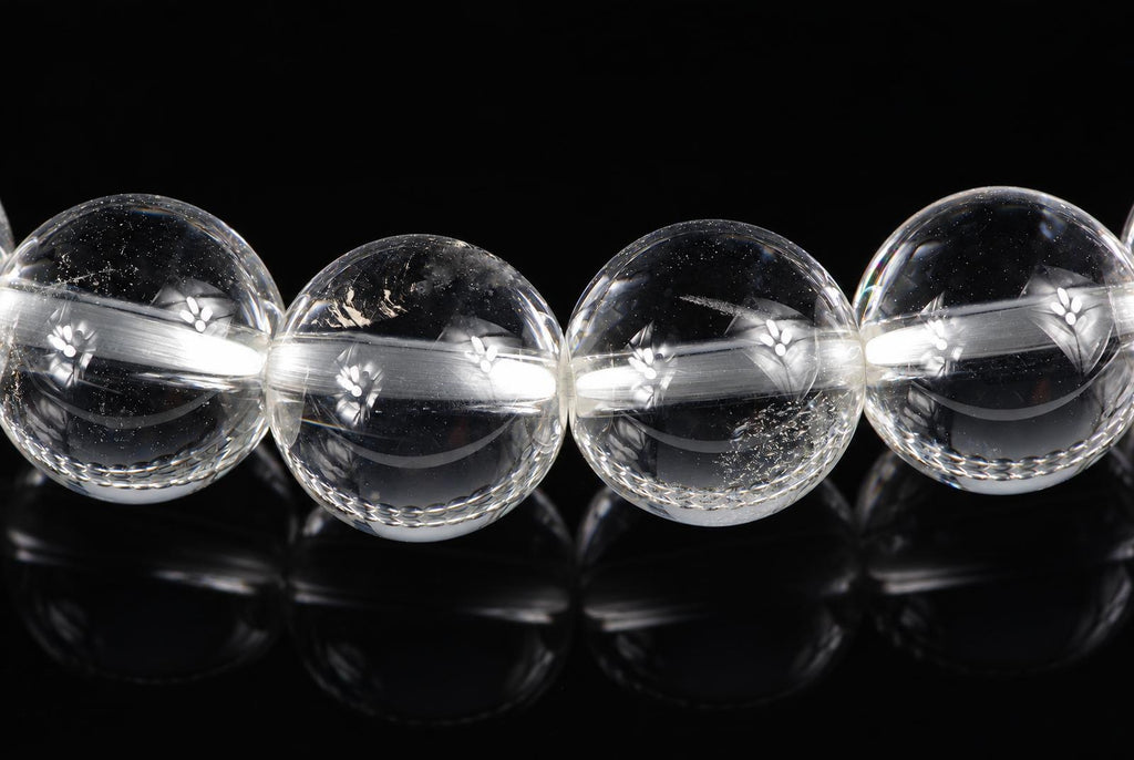 Four clear quartz beads with engraved floral patterns, arranged in a row on a dark reflective surface.