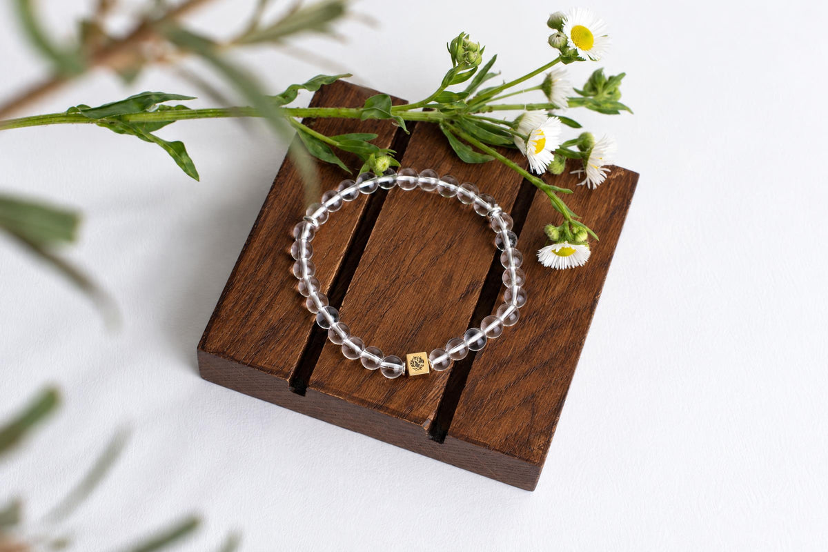 A bracelet with clear round beads and a small gold-coloured square charm rests on a dark wooden surface, surrounded by white flowers.