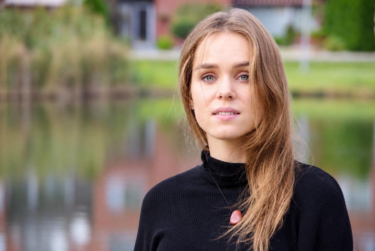 A woman with long light brown hair wears a black turtleneck and a necklace with a smooth, reddish-brown pendant against a blurred outdoor background.